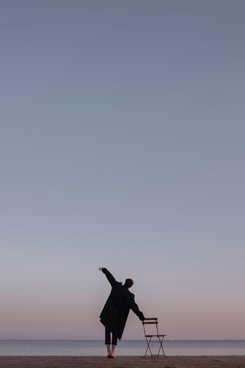 A man stands barefoot on the beach, balancing with a chair against a picturesque sunset sky.