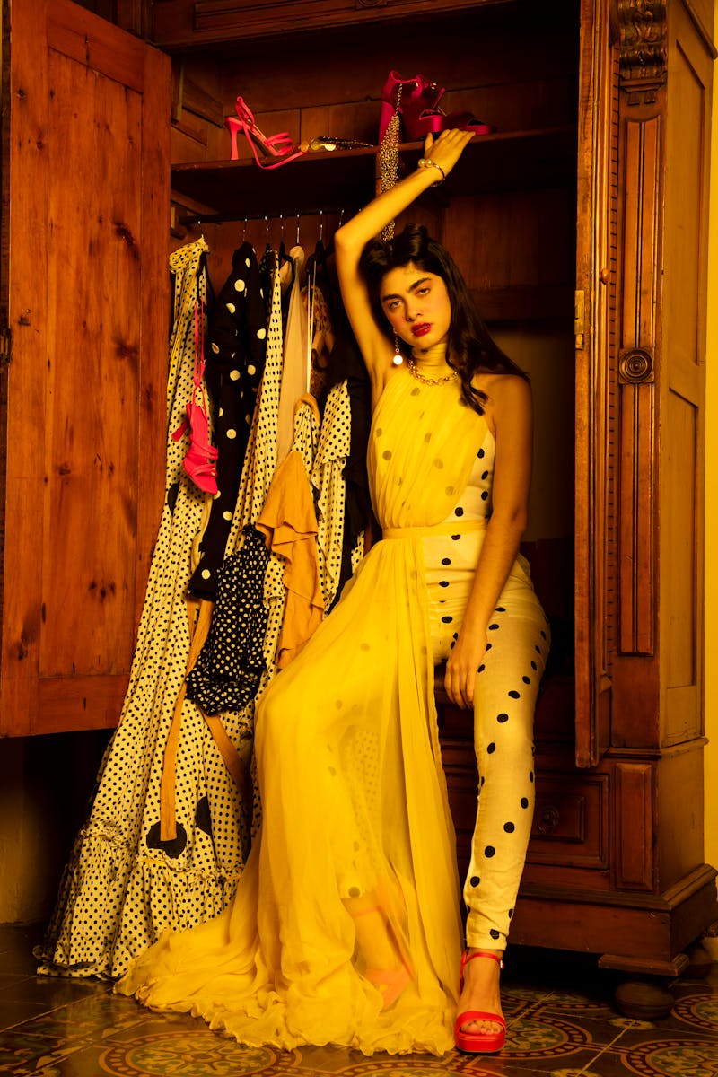 Stylish woman posing in yellow dress by wooden wardrobe showcasing fashion.