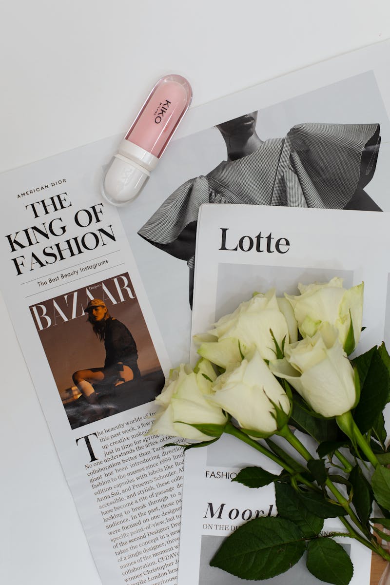 Aesthetic flat lay of fashion magazines with white roses and beauty product, conveying elegance.