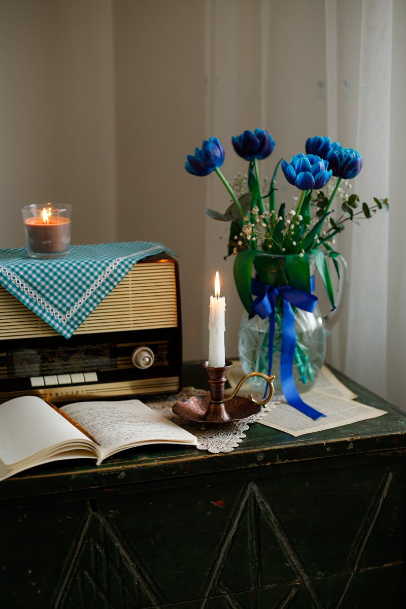 A cozy vintage setup featuring a retro radio, candle, open book, and blue flowers in a vase. Perfect for a nostalgic ambiance.