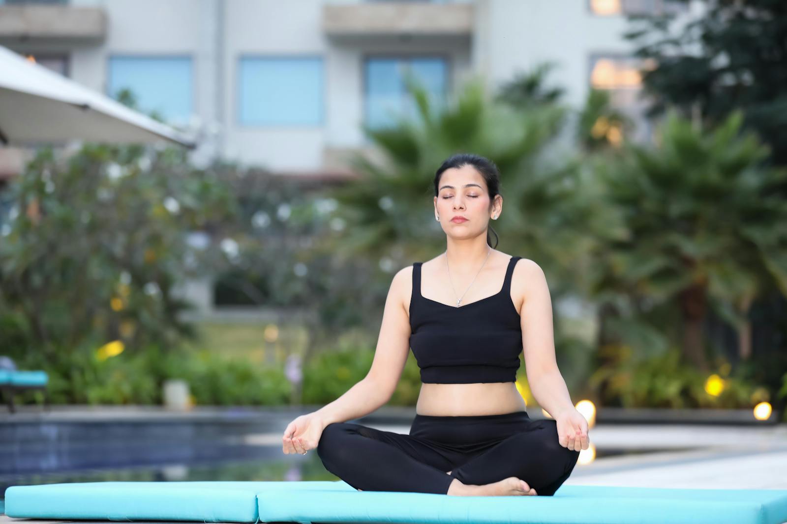A woman in black activewear meditating outdoors on a yoga mat by the poolside.