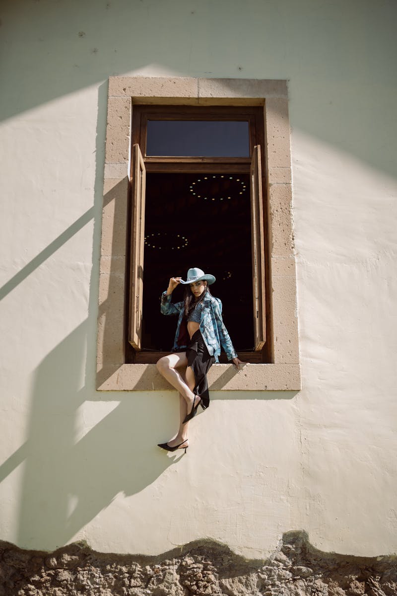 A woman in cowgirl attire sitting on a window frame of a rustic building, casting a shadow.