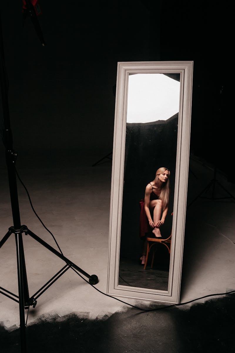 Elegantly posed woman sitting and reflected in a full-length mirror in a dark studio setting.