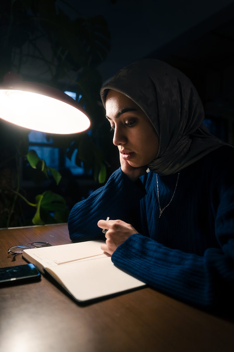 A woman in a hijab studies under a lamp, notebook open, creating a focused atmosphere.