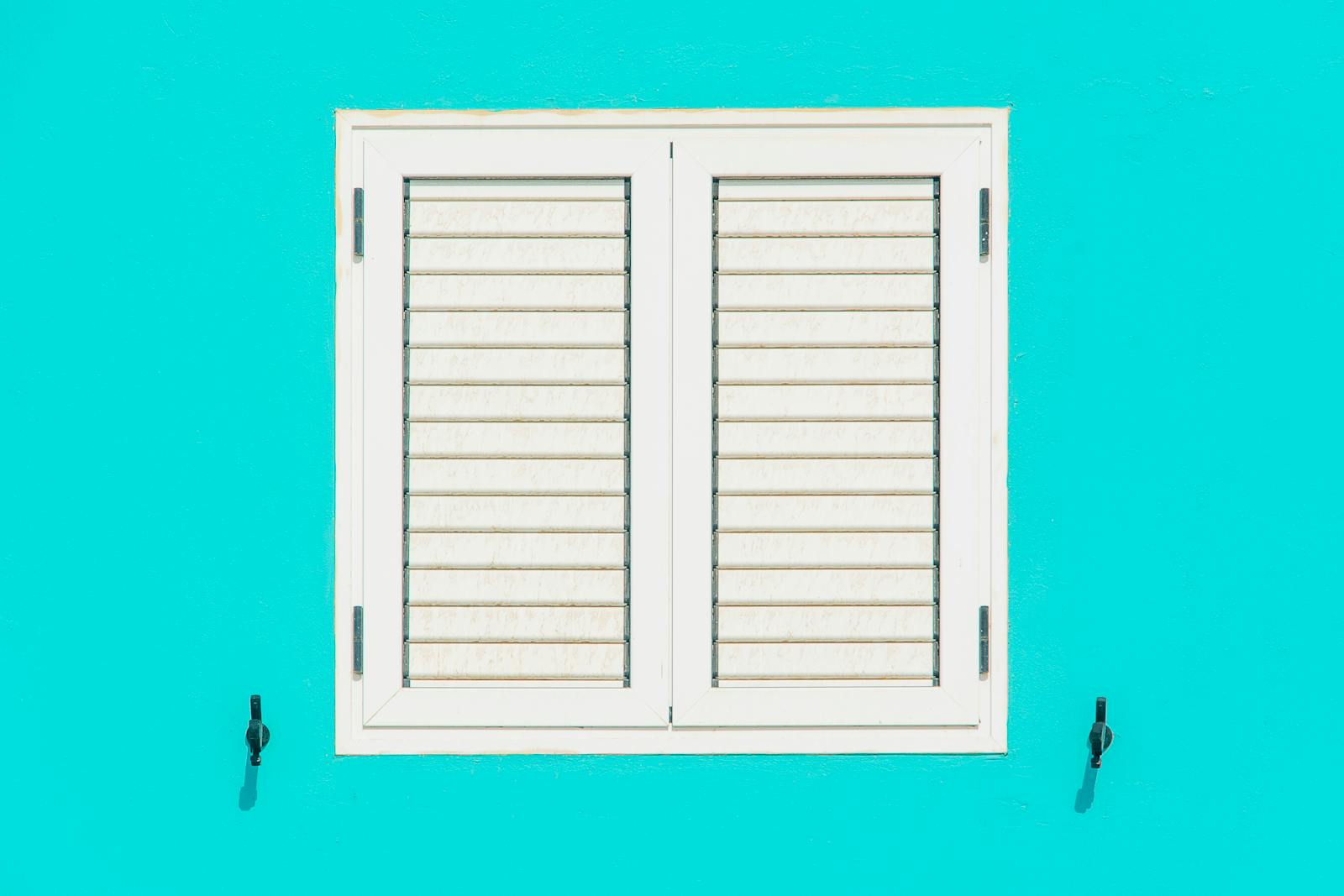 A white shuttered window set on a bright turquoise wall, exuding minimalist charm.