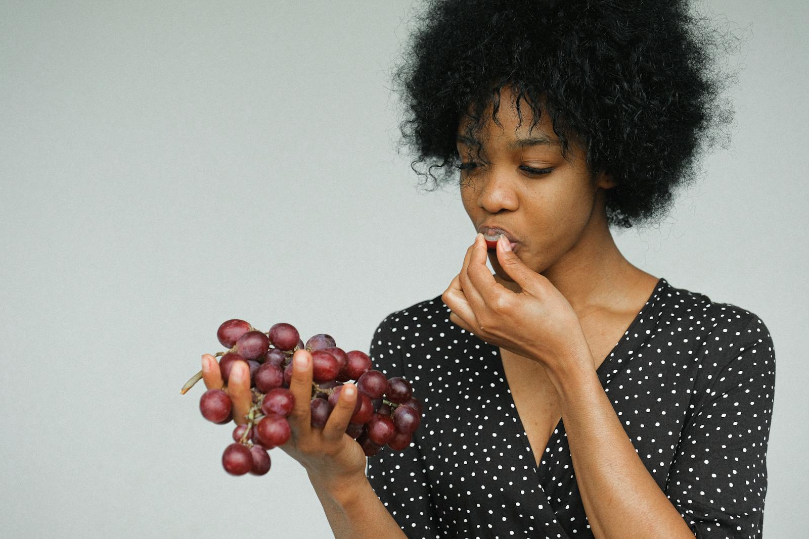 Young woman enjoying grapes against a neutral background, emphasizing simplicity and elegance.