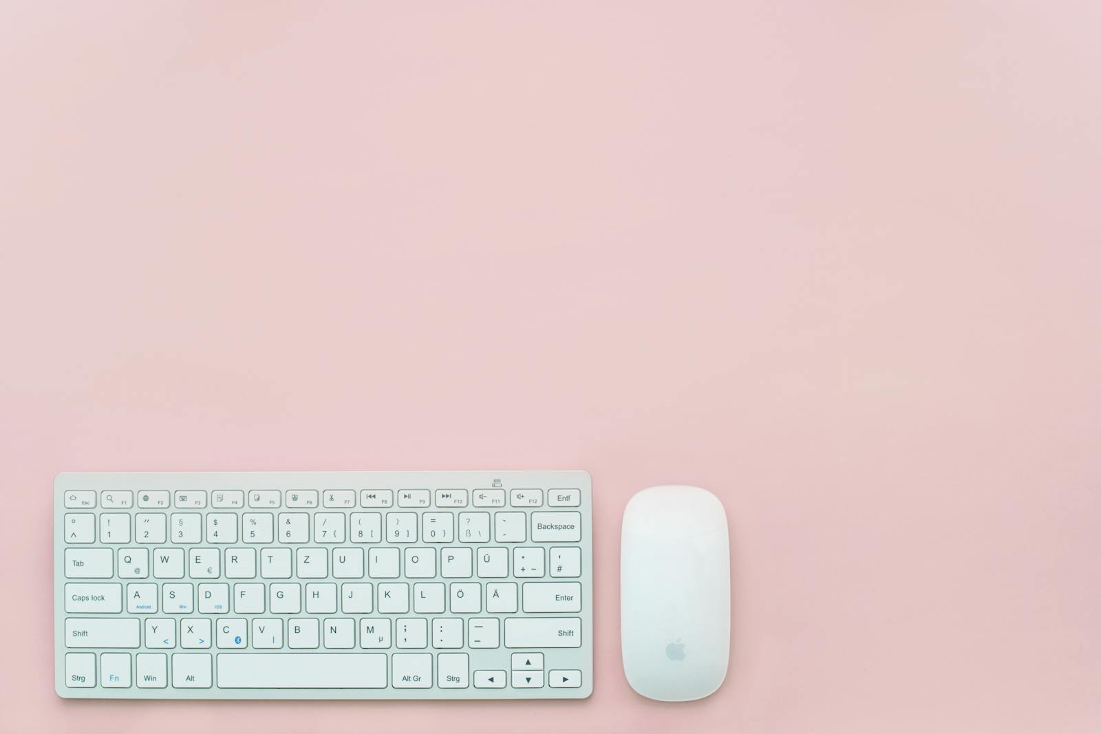 A clean and minimalist flat lay of a white keyboard and mouse on a pastel pink background. Perfect for feminine office themes.