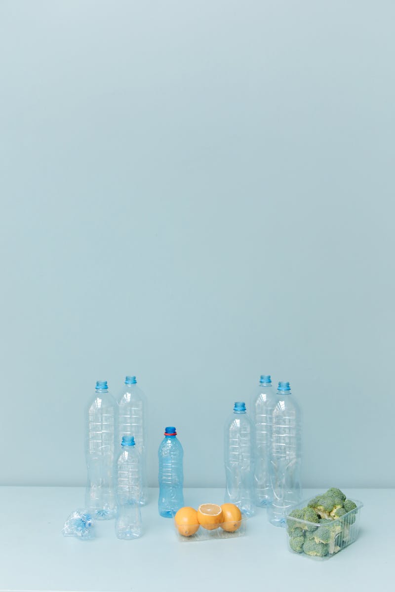Plastic bottles with broccoli and lemons on a pastel blue surface, symbolizing sustainability.