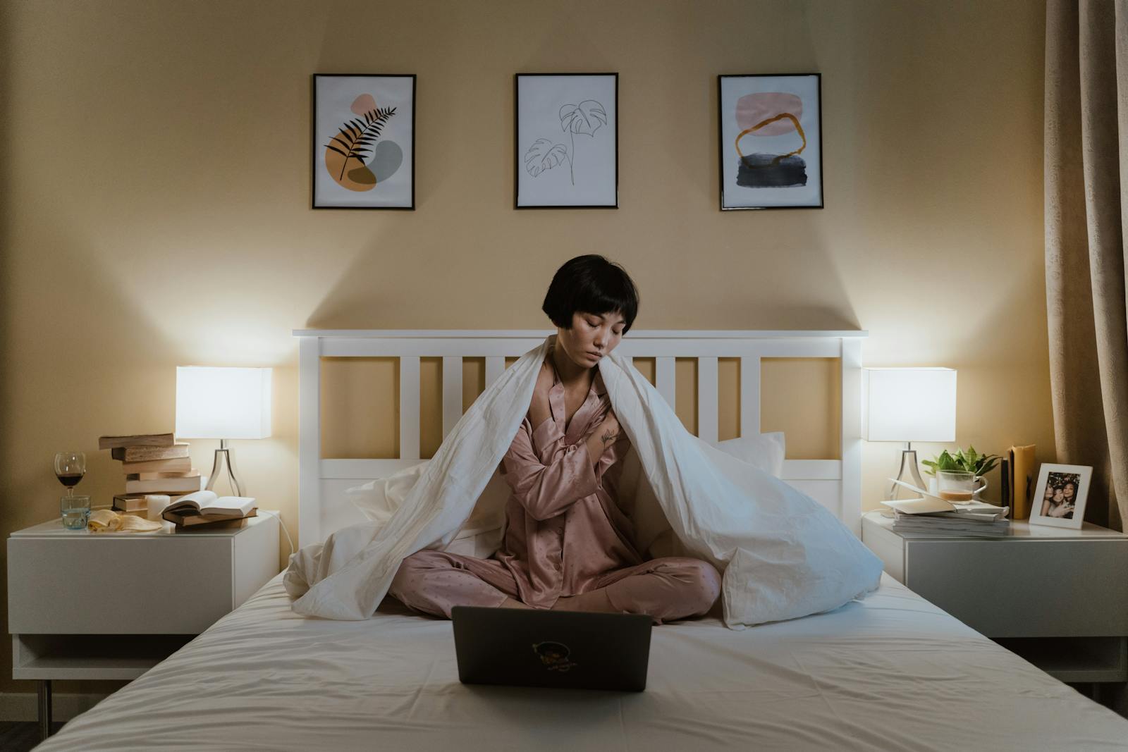 A woman sits thoughtfully in bed with a laptop amidst evening lighting.