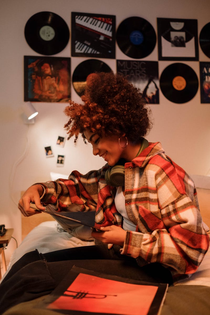 A woman with curly hair sitting on a bed enjoying a music vinyl collection indoors.
