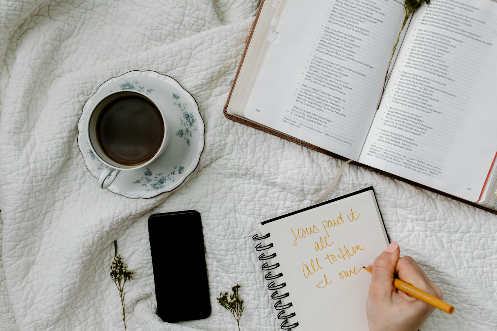 Flat lay of Bible study with coffee, notebook, and phone on a quilted blanket.
